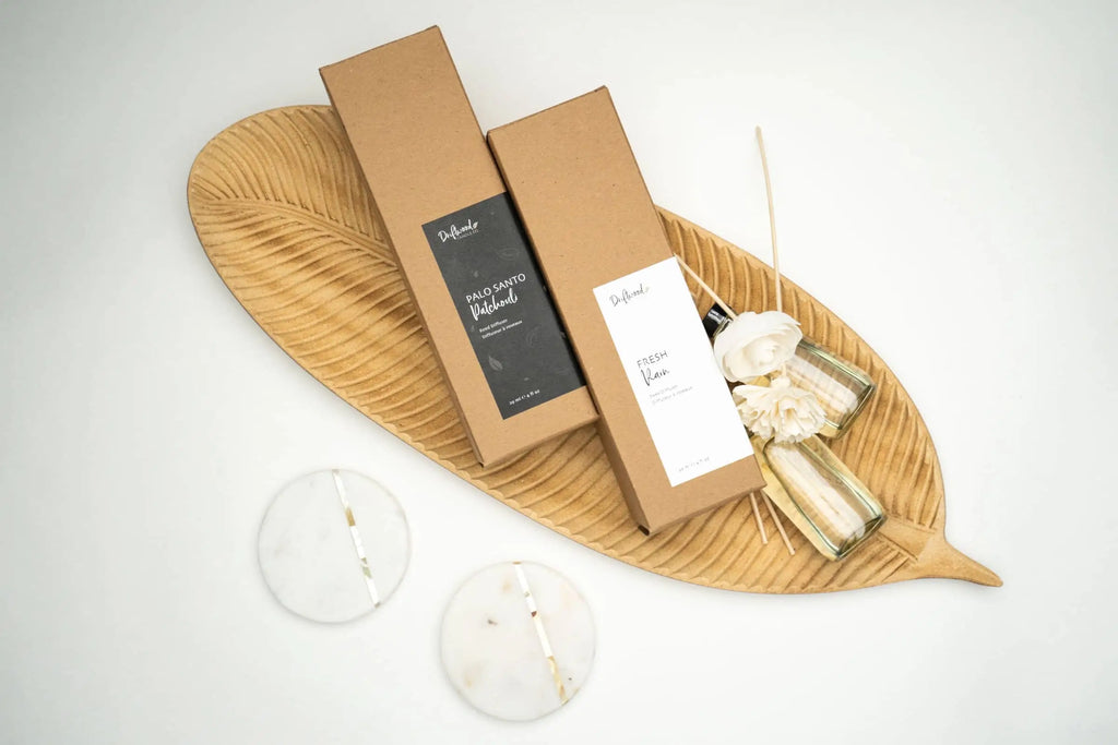 Overhead shot of two sola flower reed diffuser set by Driftwood Candle Co. lying sideways atop a decorative wooden leaf tray.  Two sola flowers are on display next to the boxes along with samples of the fragrance oil bottles.