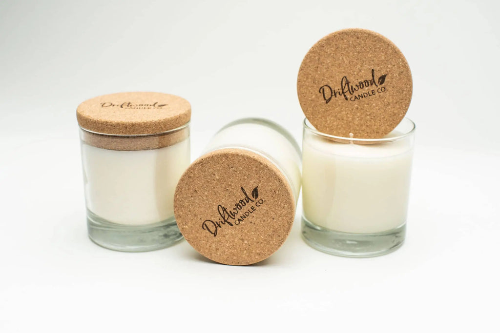 Three soy candles.  First is upright with cork lid on it.  The second is sitting on its side with branded Driftwood Candle Co. logo on cork lid, the third is upright with a cork lid resting on top on its side.