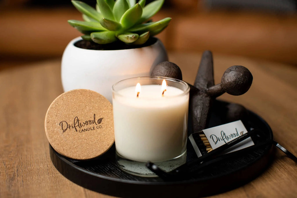 Lifestyle shot of double wick Driftwood Candle Co. candle in front of a succulent plant on a round black decorative coffee table saucer.  Stamped cork lid with logo branding is resting next to the candle along with a wick trimmer and set of matches.