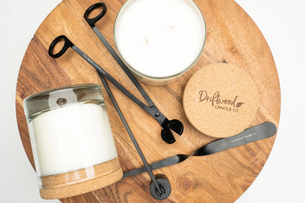Overhead shot of two candles on a round wooden tray with candle care kit randomly displayed with a branded Driftwood Candle co. cork lid next to them.