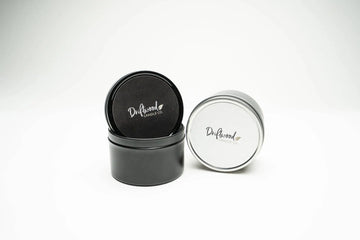 Two travel tin candles set on a white backdrop.  Tin on the left is black with lid balancing on top displaying affixed label.  Second tin is silver on it's side with labelled lid facing.