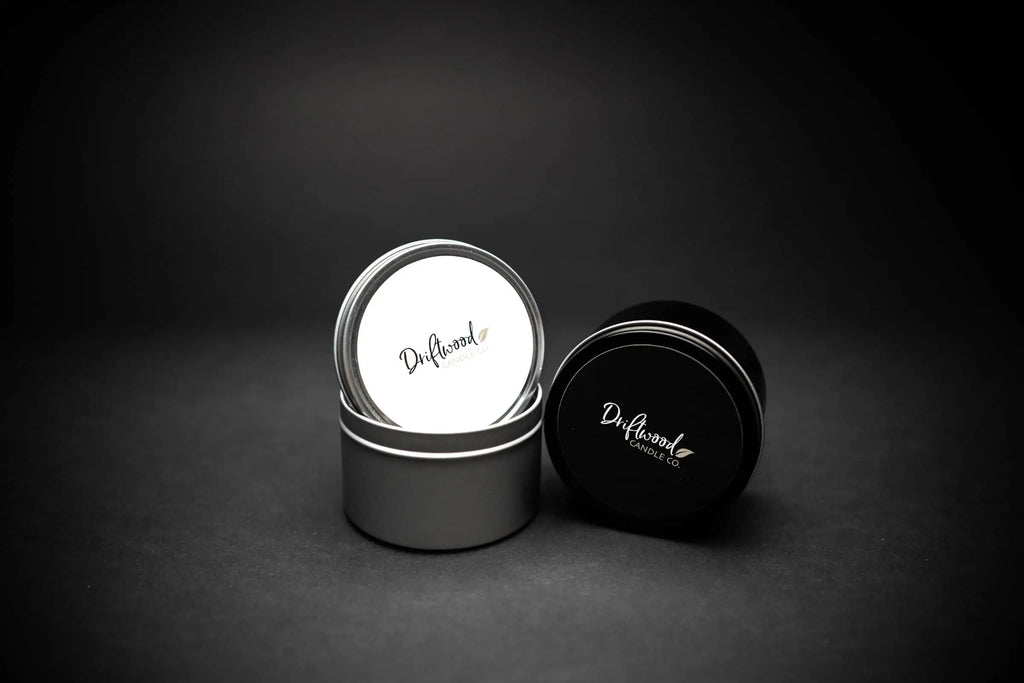 Two travel tins set on a black back drop.  One tin is silver with the lid balancing on the top with the Driftwood Candle Co. logo displayed, the other is a black tin on it's side with same label in black.