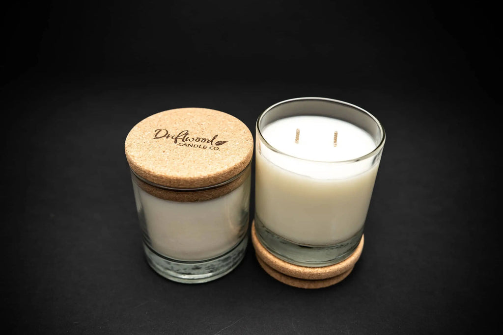 Two Driftwood Candle Co. scented soy candles on a black backdrop.  One double wick candle is nestles atop the cork lid, the other candle next to has the lid secured on top demonstrating how to use the lid when the candle is not in use.