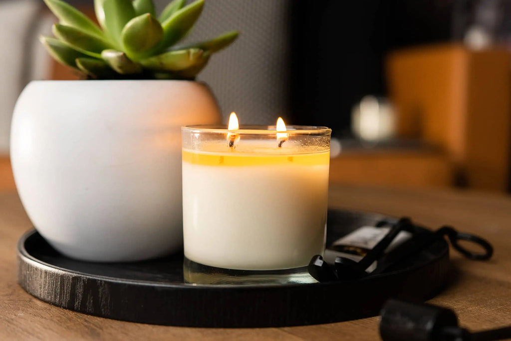 Double wick candle lit on display next to a beautiful plant.  Sitting on a round wood tray with wick trimmers and matches.