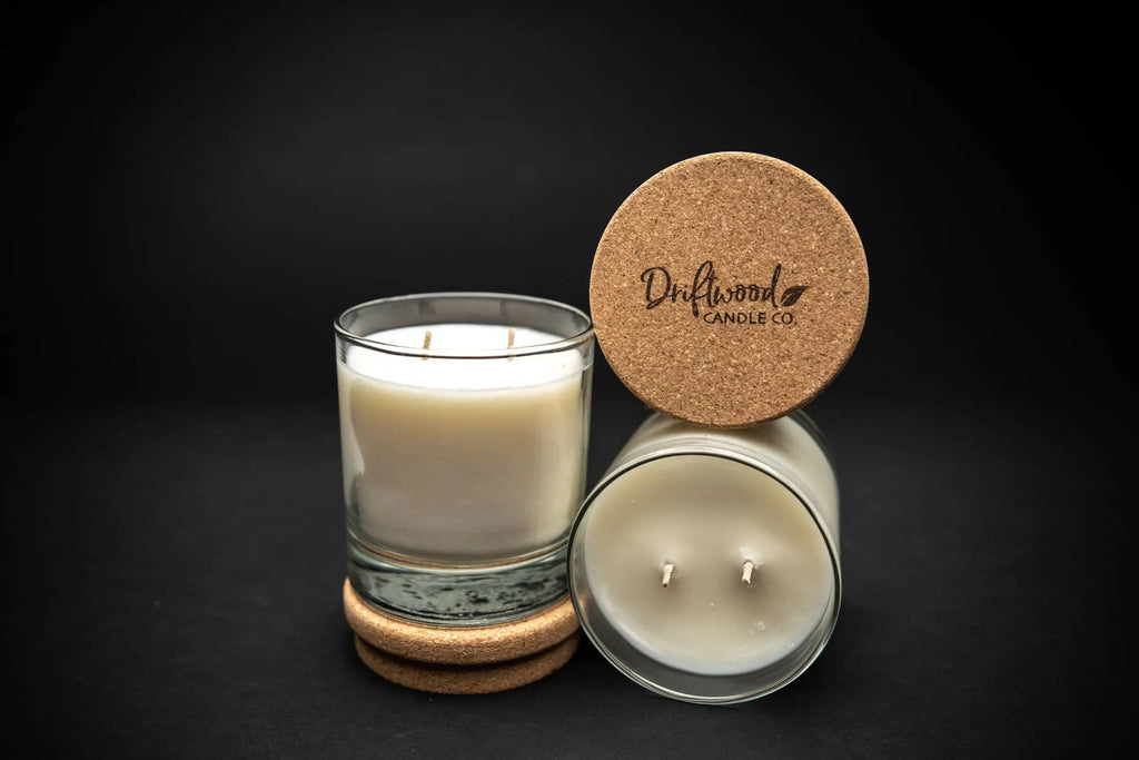 Two double wick soy candles made by Driftwood Candle Co. Scented with Amber Tobacco fragrance.  One jar is nestled on a cork lid, the other lid is balanced on top of the scented candle.