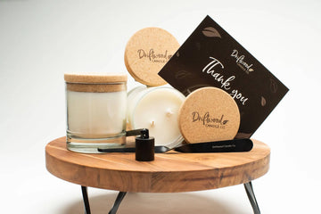 Two Driftwood Candle Co. scented soy candles on display on a woodent table riser adorned with samples of the cork lids with label on them, a candle snuffer and a candle dipper along with the thank you card on display.