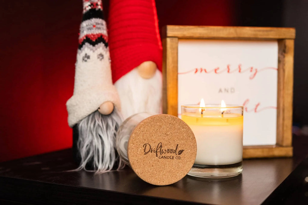 Double Wick Soy Holiday Candle burning.  Soy wax has a beautiful melt pool.  Two Christmas gnomes and a holiday sign are blurred in background.