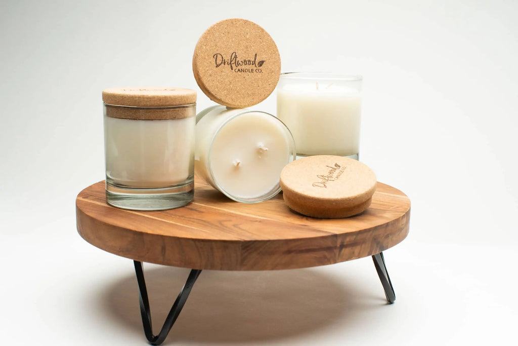 Three Driftwood Candle Co. reusable whiskey glasses filled with double wick amber tobacco scented soy wax. Includes a cork lid stamped with the logo displayed on a lifted wooden table tray.
