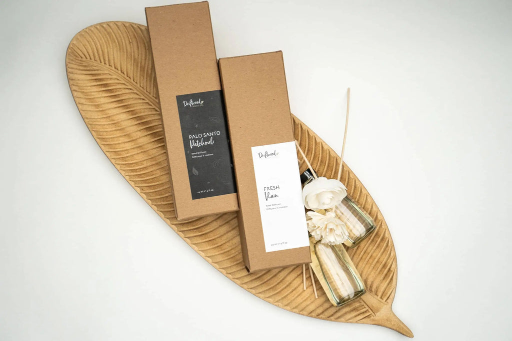 Two Driftwood Candle Co. scented reed diffuser sets on lying on display on a decorative wooden leaf tray.  Next to the boxes are two 4oz scented reed diffuser bottles filled with the fragrances along with sola flowers and reed sticks.